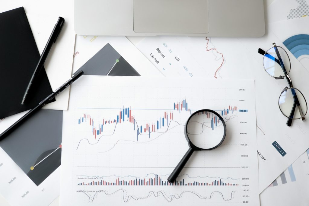 Graphiques financiers avec une loupe, ordinateur portable et lunettes sur un bureau, illustrant l’analyse et la stratégie financière.